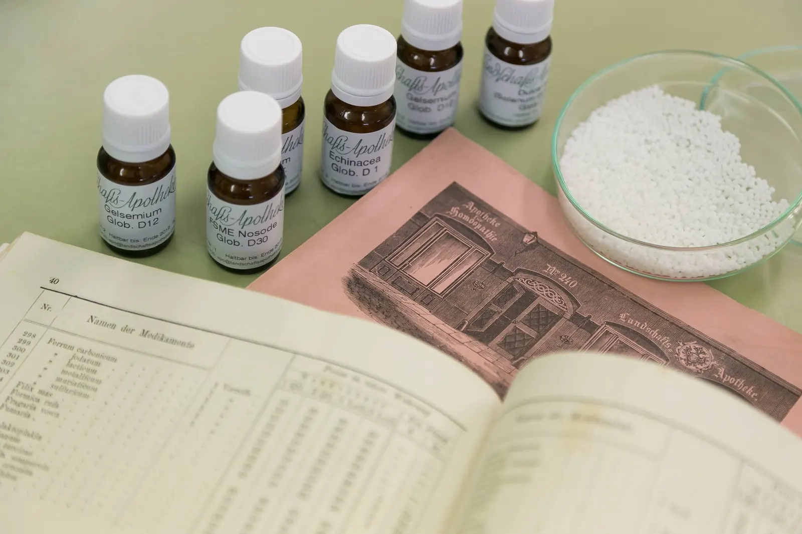 Braunglasfläschchen mit homöopathischen Arzneien, eine Schale Globuli und ein aufgeschlagenes altes Rezeptbuch